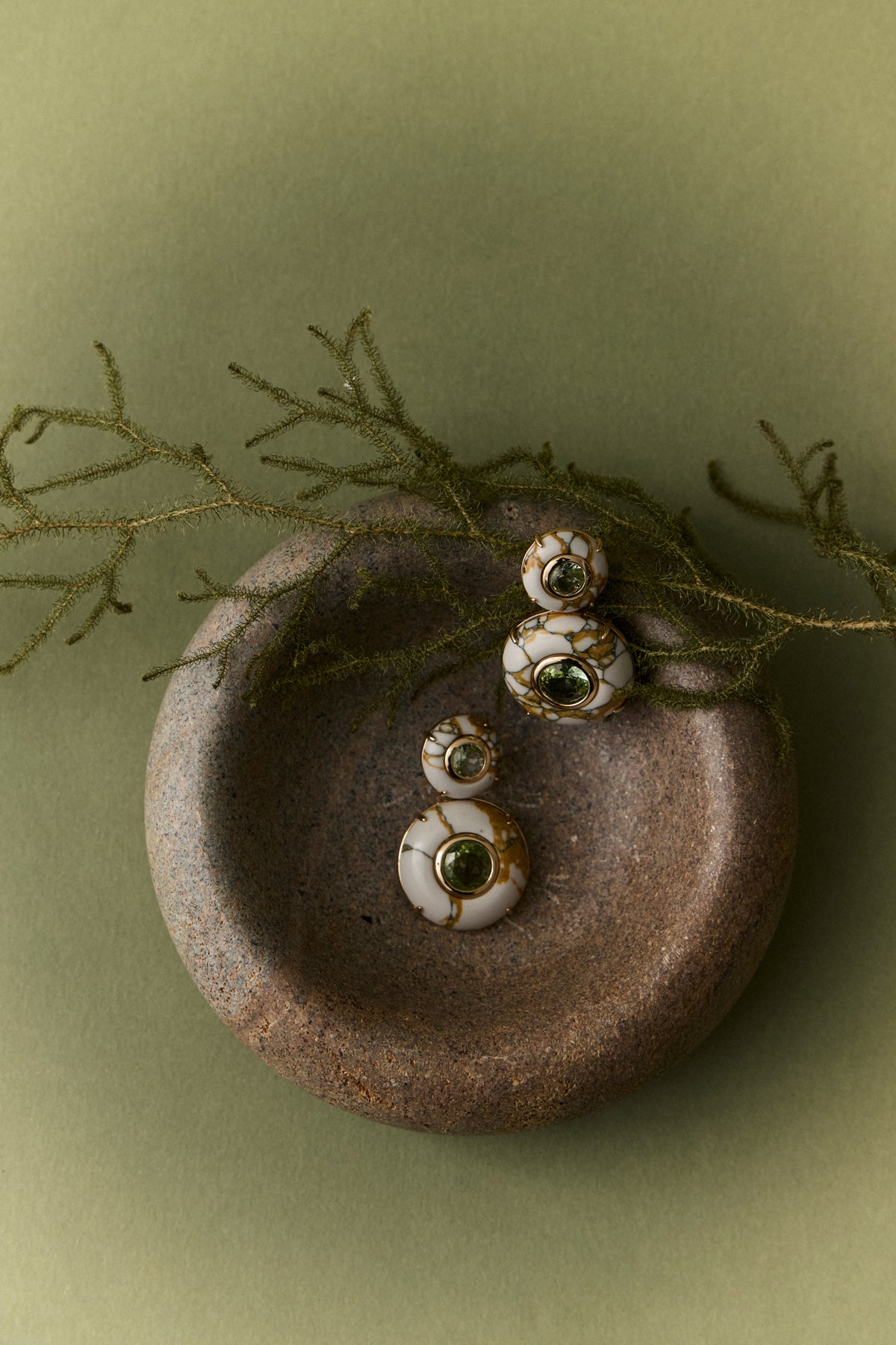BALBINO LITHOS earrings arranged on a rounded vessel beside botanical stems against a muted green background.